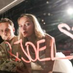 Eine Frau und ein Mann, dem der Mund verklebt ist, sehen in einem Spiegel auf dem Leyla und Joel steht.